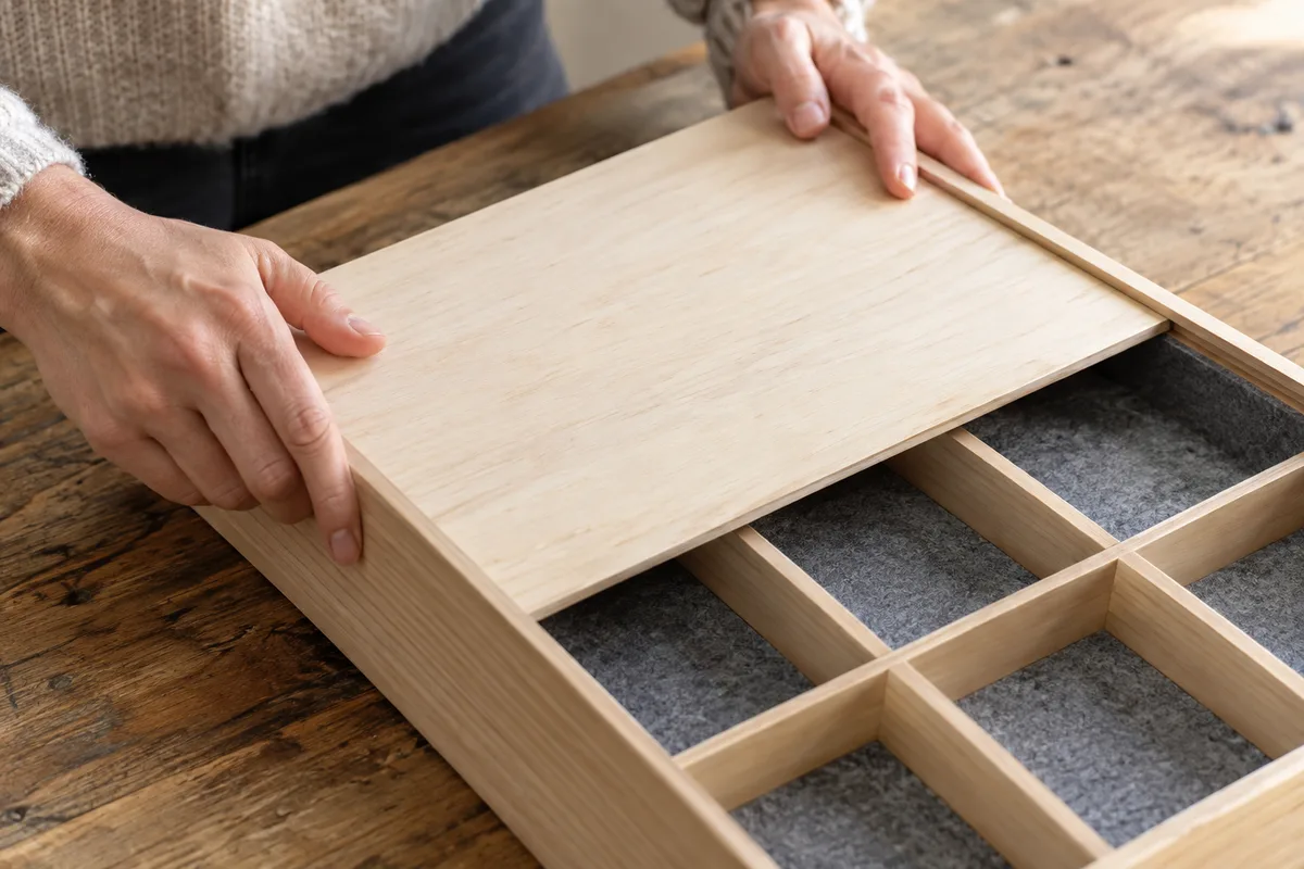 Mains d’adulte qui font glisser un couvercle en bois clair sur un coffret compartimenté posé sur une table en bois